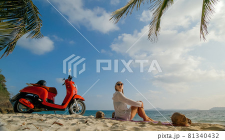 Scooter road trip. Woman alone on red motorbike in white clothes on sand beach by ocean. One girl caucasian tourist walk near tropical palm tree. Scooter road trip. Woman alone on red motorbike in white clothes on sand beach by ocean. One girl caucasian tourist walk near tropical palm tree. 81340432