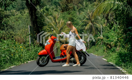 Woman on red scooter in white clothes drive on forest road trail. Dancing trip. One girl caucasian tourist in sunglasses dance, relax near motorbike. 81340446