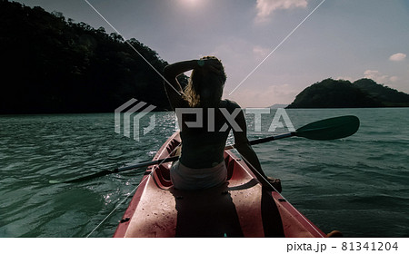 Silhouette of woman tourist raising hands while sailing in canoe along sea bay water to distant hilly island backside view. Silhouette of woman tourist raising hands while sailing in canoe along sea bay water to distant hilly island backside view. 81341204