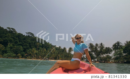 Pretty young blonde woman in stylish swimsuit and sunglasses sits on pink canoe sailing along sea against hills. 81341330