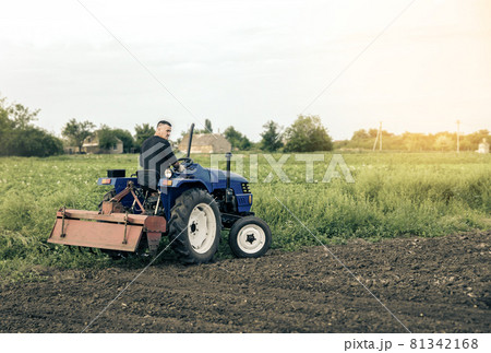 A farmer is driving a tractor across the field. Mill grinding machine for soil. Land cultivation. Recruiting and hiring seasonal worker for work. Farming, agriculture. Land improvement, mitigation. 81342168