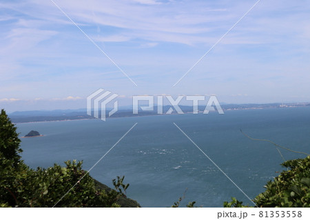 緑の樹々の向こう見える丸い小さな島のある白波のたつ青い海と少し霞んで見える対岸の山々と街と薄雲と青空 緑の樹々の向こう見える丸い小さな島のある白波のたつ青い海と少し霞んで見える対岸の山々と街と薄雲と青空 81353558