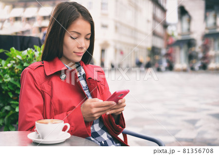 Cafe woman using mobile phone drinking cappuccino coffee at outdoor street european city. Europe travel lifestyle. Asian businesswoman in fashion red trench coat 81356708