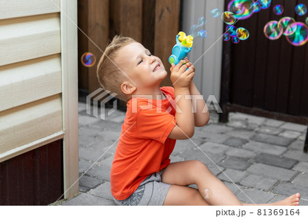 Portrait of cute little bond kid boy enjoy having fun play blowing soap bubbles siiting at home yard outdoors on bright warm summer day against house wall. Child healthy outside nature activities 81369164