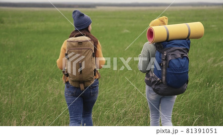 Happy active hipster girls with backpacks on their backs travel through a beautiful valley, a millennial woman on a green field, travel hiking, hiking on the trail, outdoor recreation 81391051
