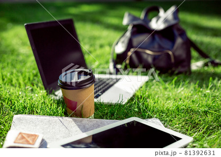 Laptop computer on green grass with coffee cup, bag and tablet in outdoor park 81392631