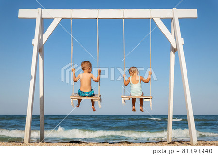 Back view couple of cute little adorable small caucasian sibling kids enjoy having fun swinging and playing at playground sea sand beach against blue ocean water wave and clear sky. Family vacation 81393940