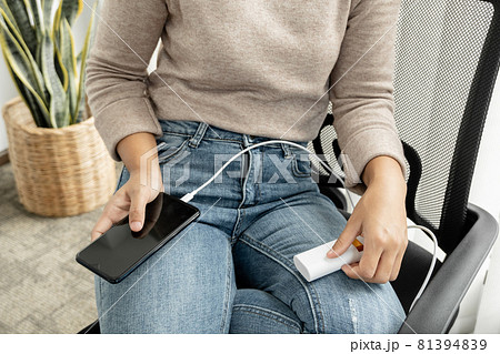 The hand of a young woman holding a smart phone and a modern spare battery charger Place the lap to charge the smartphone with a spare battery charger and play on a chair in room 81394839