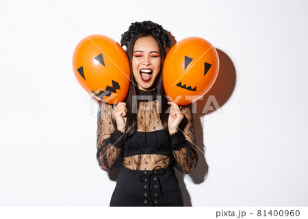 Image of asian girl in evil witch costume holding two orange balloons with scary faces, celebrating halloween, standing over white background 81400960