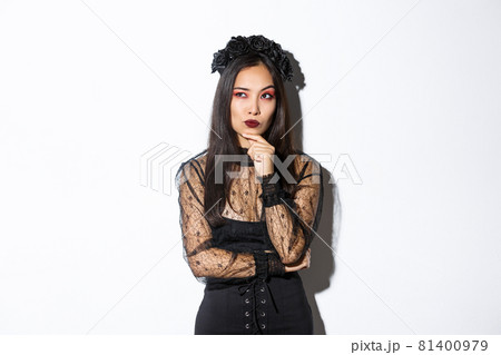 Thoughtful elegant evil witch in black lace dress, gothic wreath and makeup looking upper left corner. Woman making plans for halloween party, thinking over white background 81400979