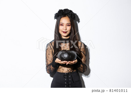 Smiling devious asian girl in witch costume, celebrating halloween, holding black pumpkin, standing over white background Smiling devious asian girl in witch costume, celebrating halloween, holding black pumpkin, standing over white background 81401011
