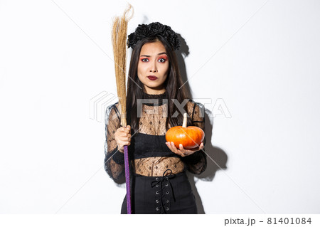 Worried and confused asian woman in witch costume looking nervous, holding broom and pumpkin, trick or treating on halloween, standing over white background 81401084