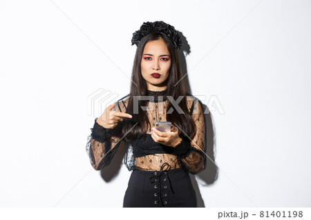 Disappointed asian woman in halloween costume, looking displeased as pointing finger at mobile phone and frowning, standing over white background Disappointed asian woman in halloween costume, looking displeased as pointing finger at mobile phone and frowning, standing over white background 81401198