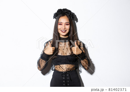 Image of happy asian woman in halloween party dress showing thumbs-up in approval. Girl wearing as witch or widow looking pleased, like or agree with something, white background 81402371