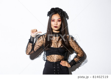 Confident stylish asian woman looking determined, wearing black lace dress for halloween party, pointing at herself sassy, standing over white background Confident stylish asian woman looking determined, wearing black lace dress for halloween party, pointing at herself sassy, standing over white background 81402519