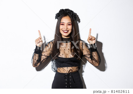 Cheerful elegant asian woman in black gothic dress and wreath, smiling happy and showing your logo, pointing fingers up at something about halloween, standing over white background Cheerful elegant asian woman in black gothic dress and wreath, smiling happy and showing your logo, pointing fingers up at something about halloween, standing over white background 81402611