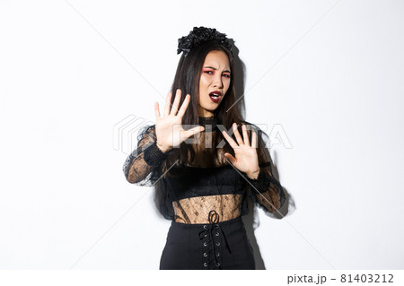 Image of bothered and annoyed asian woman in elegant gothic dress raising hands defensive, grimacing from camera flesh, asking to stop taking pictures of her, standing white background Image of bothered and annoyed asian woman in elegant gothic dress raising hands defensive, grimacing from camera flesh, asking to stop taking pictures of her, standing white background 81403212