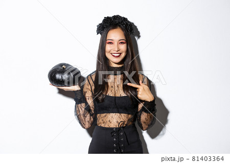 Happy smiling asian woman enjoying halloween celebration, wearing witch costume and pointing finger at black pumpkin, standing over white background Happy smiling asian woman enjoying halloween celebration, wearing witch costume and pointing finger at black pumpkin, standing over white background 81403364