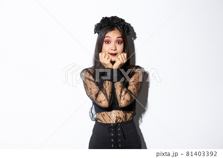 Excited cute asian girl in witch costume looking hopeful at camera, daydreaming while standing over white background, wearing gothic lace dress with wreath 81403392