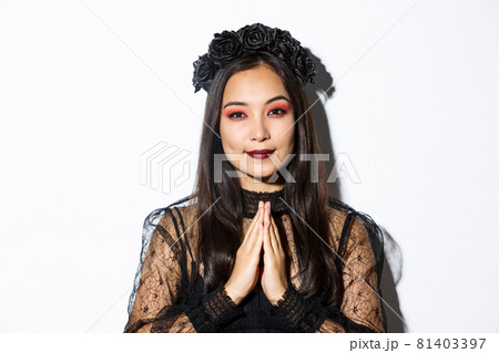 Close-up of young asian woman in black gothic dress and wreath holding hands in pray, girl wearing witch costume and celebrating halloween Close-up of young asian woman in black gothic dress and wreath holding hands in pray, girl wearing witch costume and celebrating halloween 81403397