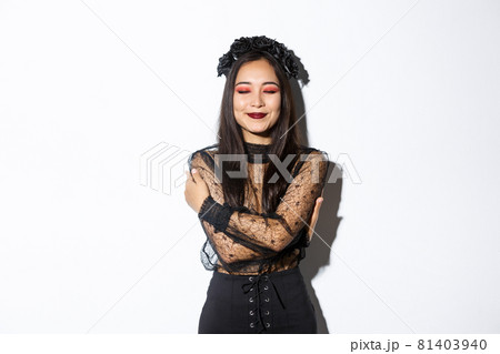 Happy cute asian woman hugging herself and smiling dreamy, wearing halloween costume of witch or widow, standing over white background Happy cute asian woman hugging herself and smiling dreamy, wearing halloween costume of witch or widow, standing over white background 81403940