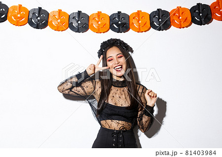 Carefree smiling asian woman in witch costume enjoying halloween party, dancing and showing peace gesture, standing over white background with pumpkin decoration Carefree smiling asian woman in witch costume enjoying halloween party, dancing and showing peace gesture, standing over white background with pumpkin decoration 81403984