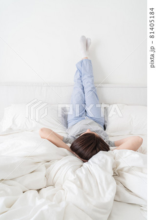 Woman wearing pajamas with legs raised up high and arms under her head lying on bed 81405184