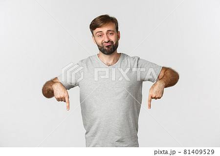 Portrait of unimpressed skeptical guy with beard, wearing gray t-shirt, sulking and pointing fingers down disappointed, standing over white background 81409529