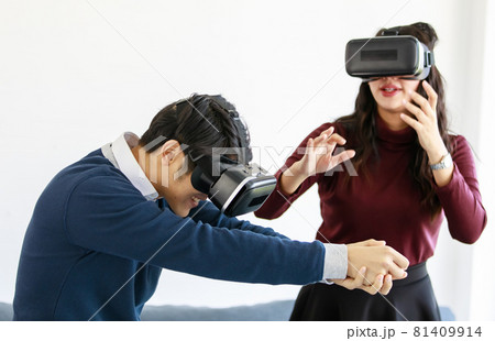 Portrait of cute smiling young Asian lover couple having fun with playing a new video game with virtual reality headset glasses together while sitting on the couch with Christmas tree background Portrait of cute smiling young Asian lover couple having fun with playing a new video game with virtual reality headset glasses together while sitting on the couch with Christmas tree background 81409914
