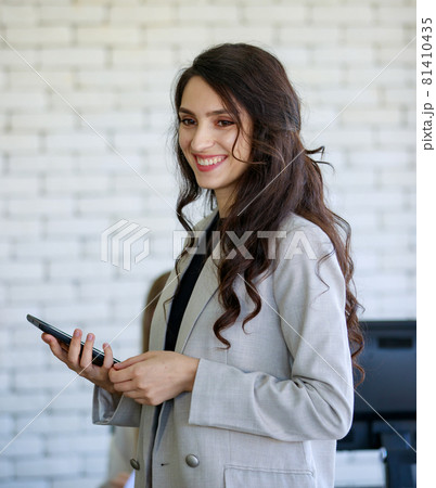 Portrait of an attractive smiling professional middle-aged businesswoman wearing gray formal clothes with dark brown hairstyle holding a digital tablet and looking to colleagues in the office 81410435