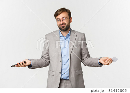 Image of clueless smiling businessman in glasses and gray suit, spread hands sideways and shrugging confused, holding mobile phone and credit card Image of clueless smiling businessman in glasses and gray suit, spread hands sideways and shrugging confused, holding mobile phone and credit card 81412006
