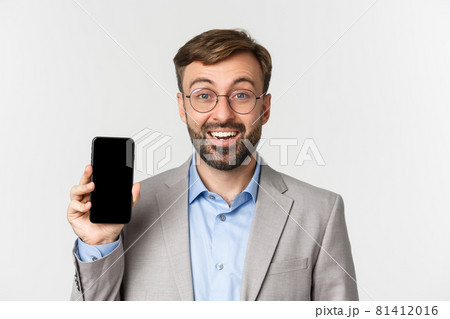 Close-up of cheerful male entrepreneur in glasses and gray suit, showing smartphone screen and smiling, standing over white background Close-up of cheerful male entrepreneur in glasses and gray suit, showing smartphone screen and smiling, standing over white background 81412016