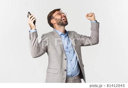Portrait of happy and relieved businessman, holding smartphone, raising hands up and shouting for joy, triumphing and celebrating victory, standing over white background Portrait of happy and relieved businessman, holding smartphone, raising hands up and shouting for joy, triumphing and celebrating victory, standing over white background 81412043