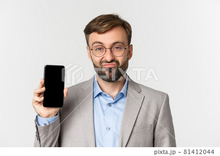 Close-up of skeptical businessman in glasses and gray suit, pouting and showing mobile phone screen, standing over white background Close-up of skeptical businessman in glasses and gray suit, pouting and showing mobile phone screen, standing over white background 81412044