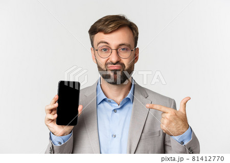 Close-up of unamused male entrepreneur in glasses and gray suit, smirking and pointing finger at mobile phone, showing something on screen 81412770