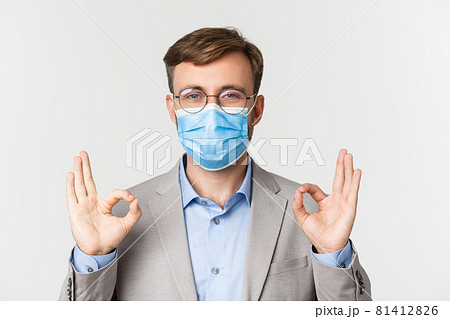 Concept of work, covid-19 and social distancing. Close-up of businessman in grey suit and medical mask, showing okay signs in approval, standing over white background Concept of work, covid-19 and social distancing. Close-up of businessman in grey suit and medical mask, showing okay signs in approval, standing over white background 81412826