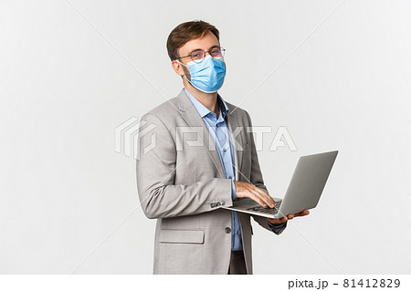 Concept of work, covid-19 and social distancing. Image of working businessman in medical mask and suit, using laptop and smiling happy, standing over white background Concept of work, covid-19 and social distancing. Image of working businessman in medical mask and suit, using laptop and smiling happy, standing over white background 81412829