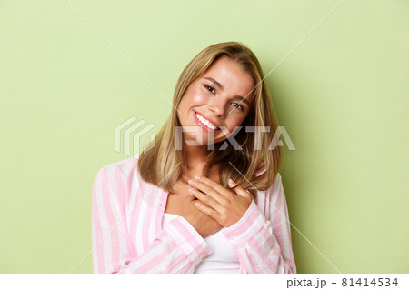 Close-up of silly blond girl in pink shirt, holding hands on heart and smiling grateful, express gratitude, standing over green background 81414534