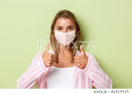 Concept of quarantine, coronavirus and lifestyle. Close-up of confident attractive girl in face mask, showing thumbs-up in approval, like something, standing over green background 81414535