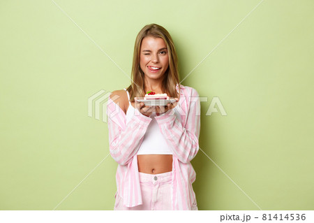 Portrait of beautiful glamour girl with blond hair, looking pleased, holding piece of cake and licking lips, standing over green background 81414536
