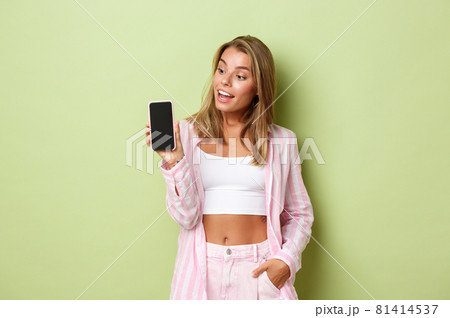 Image of blond attractive female model in pink shirt, showing smartphone screen and looking amazed, standing over green background 81414537