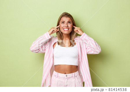 Portrait of annoyed woman complaining on loud neighbours, looking up and frowning, shut ears with fingers, standing over green background 81414538