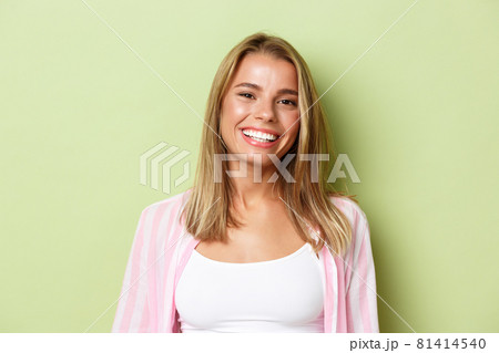 Close-up of stylish blond girl smiling, looking confident, standing over green background 81414540