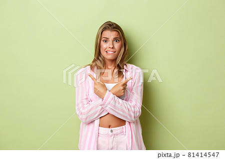 Portrait of worried indecisive woman with blond short hairstyle, wearing pink outfit , looking nervous while pointing fingers sideways at two variants, green background 81414547