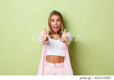 Image of attractive happy female model with blond hair, wearing pink shirt and jeans, showing thumbs-up in approval, like something, standing over green background Image of attractive happy female model with blond hair, wearing pink shirt and jeans, showing thumbs-up in approval, like something, standing over green background 81415995