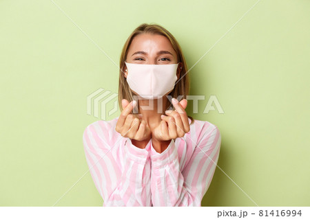 Concept of quarantine, coronavirus and lifestyle. Close-up of beautiful blond female model in face mask and pink shirt, showing heart sign, standing over green background 81416994