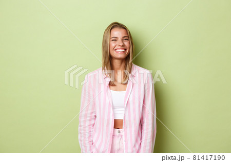 Image of cheerful blond woman with white perfect smile, standing over green background happy, wearing pink outfit Image of cheerful blond woman with white perfect smile, standing over green background happy, wearing pink outfit 81417190