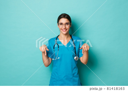 Coronavirus, pandemic and social distancing concept. Image of attractive female doctor in scrubs, pointing fingers down and smiling, showing logo, standing over blue background 81418163