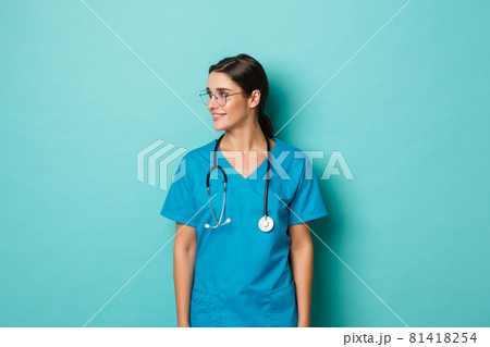 Coronavirus, pandemic and social distancing concept. Close-up of beautiful female doctor in glasses and scrubs, looking left and smiling at copy space, standing over blue background 81418254