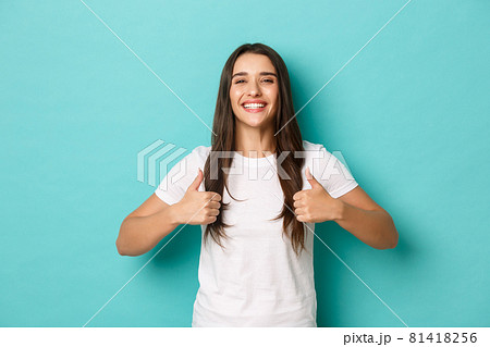 Image of satisfied smiling brunette girl, wearing white t-shirt, showing thumbs-up in approval, like and recommend product, standing over blue background 81418256